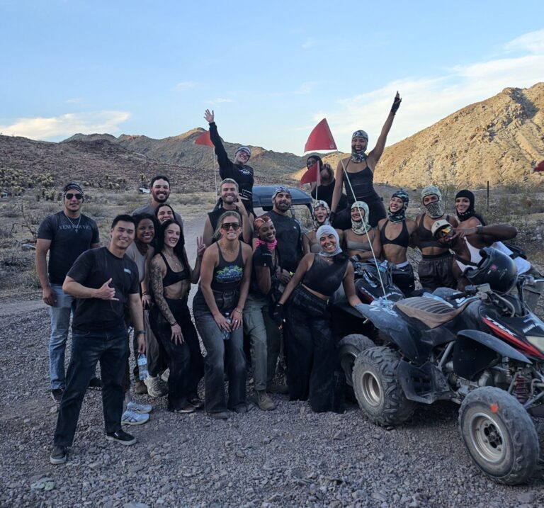 Group enjoys ATV tour near Las Vegas, Nevada, posing on rocky desert terrain with mountains and clear blue sky in the background.
