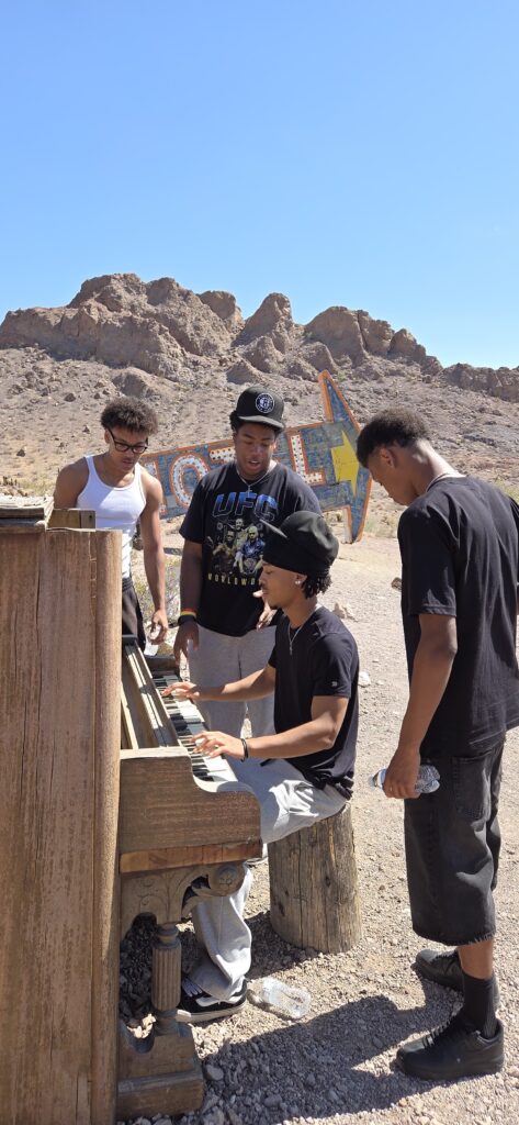 Four young men in casual wear gather at an upright piano in Nevada’s desert near a colorful sign, perfect for Ghost Town sightseeing.
