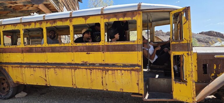 Tour group relaxes in a vintage yellow bus with missing windows near a Nevada ghost town by the Colorado River under blue skies.
