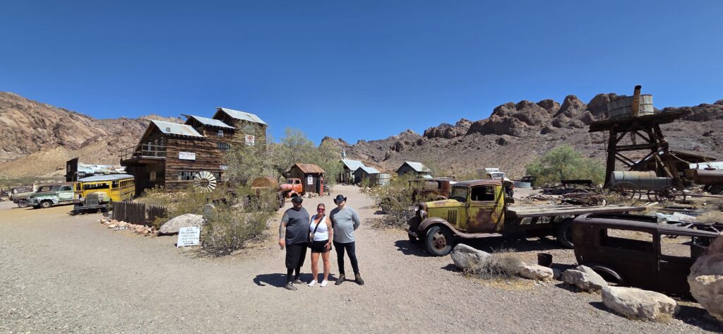 Three visitors pose by rustic buildings and vintage cars in Nelson Ghost Town, Nevada near Las Vegas—great for sightseeing or ATV tours.