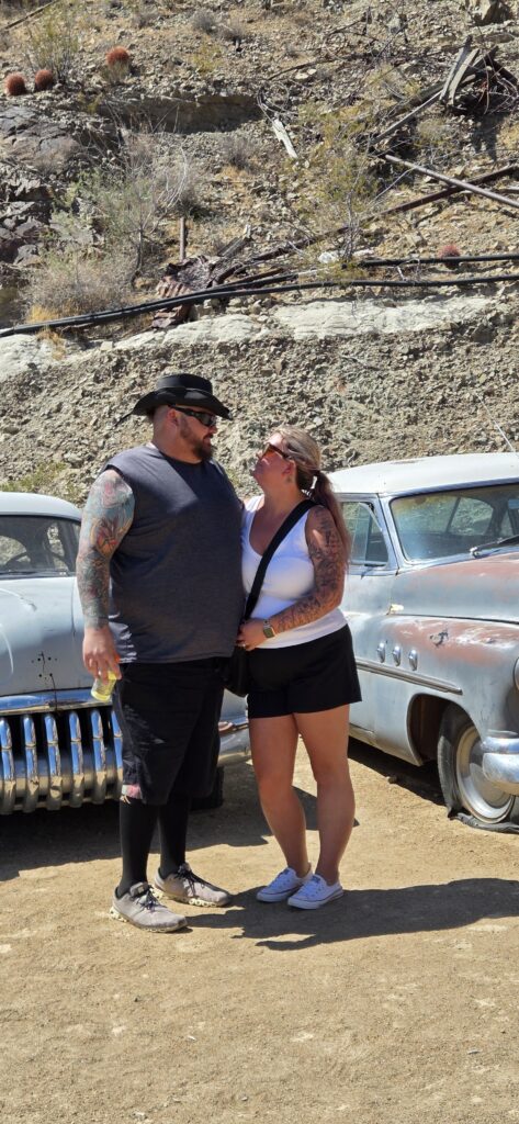 Couple by vintage cars in Nevada desert near Las Vegas, smiling with rocky hills, cacti—perfect for ATV or Ghost Town tours.