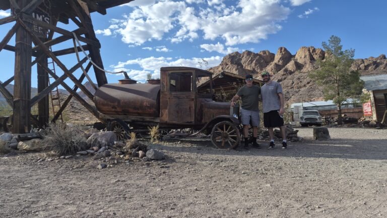 Two people by a rusty old truck in a Nevada desert ghost town near Las Vegas, with rocky hills and sparse vegetation.