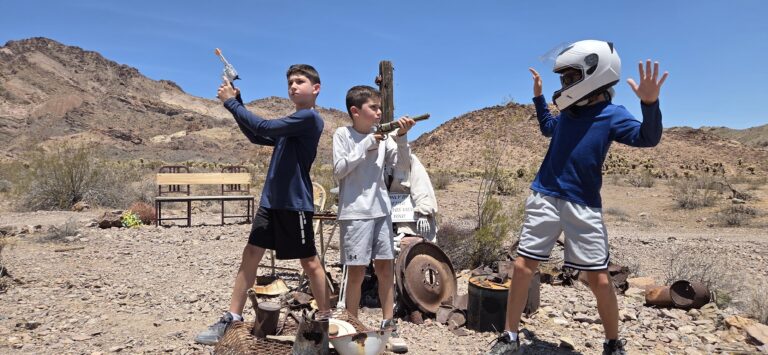 Three boys play near rocky Nevada hills by the Colorado River, with toy guns and helmets; benches and desert gear scattered around.