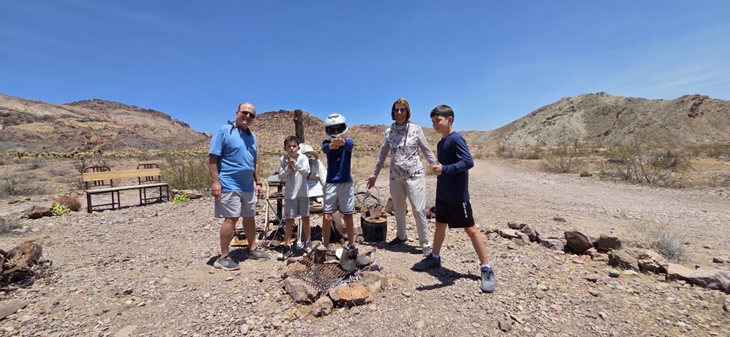 Family with kids smiles by a fire pit in a Nevada desert near Las Vegas, mountains, empty chairs, and desert plants visible.