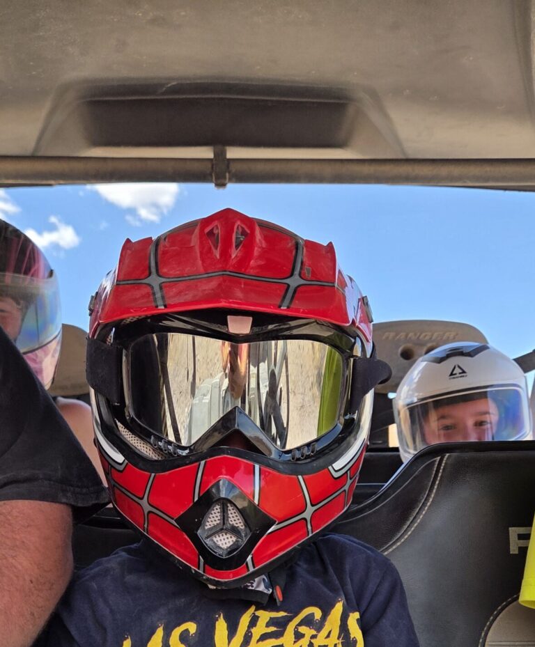 Child in red motocross gear and Las Vegas shirt on RZR off-road ride near the Colorado River, with helmeted riders in back.