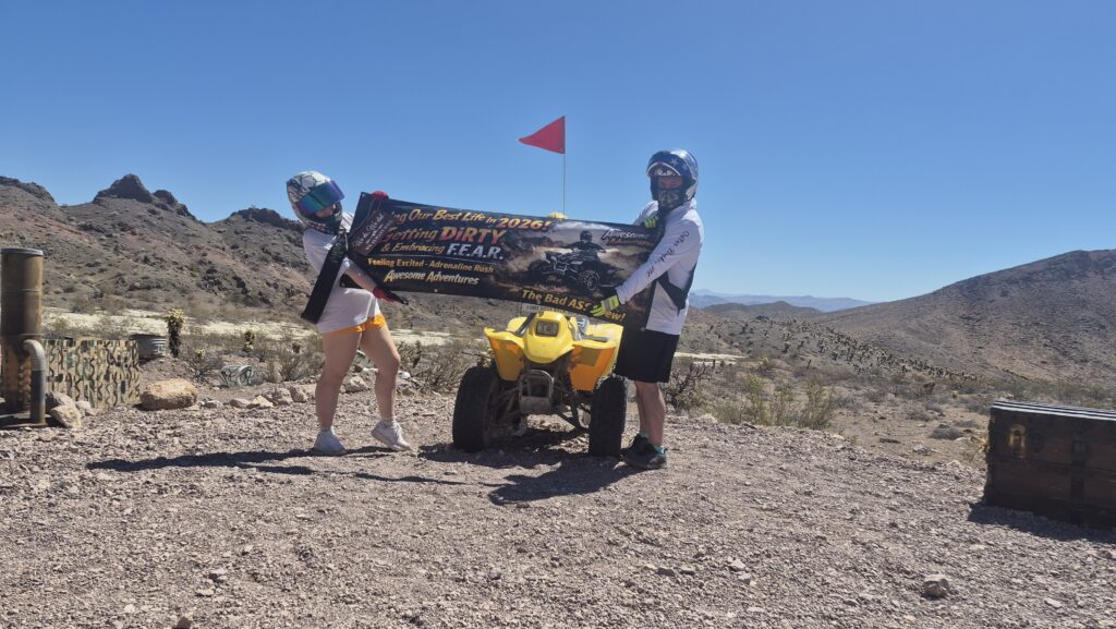 Two people in helmets on rocky Nevada terrain hold a banner by a yellow ATV, ready for Las Vegas ATV tours near desert mountains.