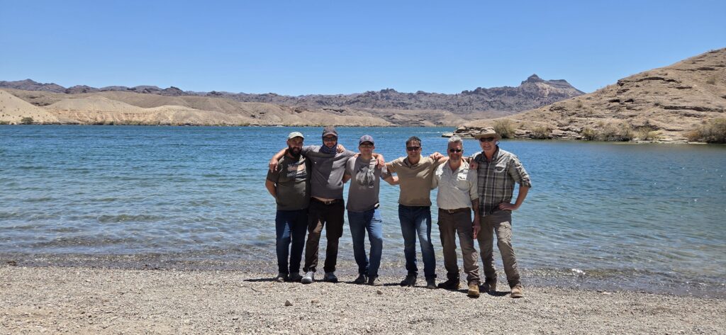 Five men smiling arm-in-arm on a pebbly Nevada shore by the Colorado River near Las Vegas, clear blue sky and hills behind.
