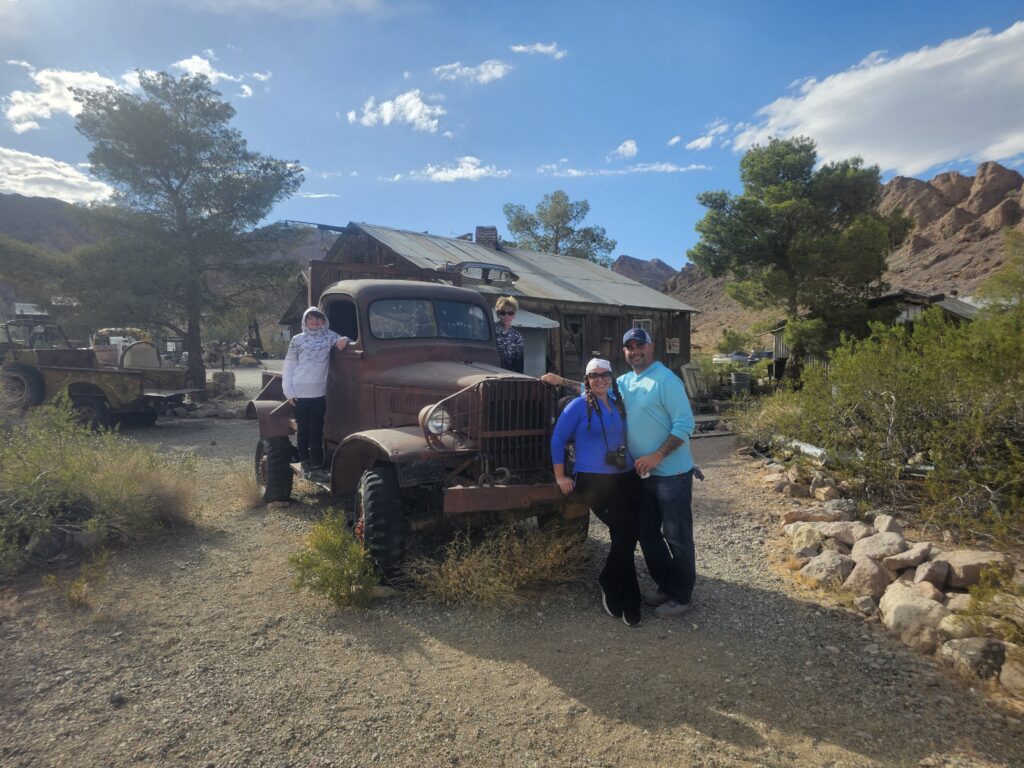 Group poses by a rusty truck at a Nevada ghost town near Las Vegas, with rocky hills—perfect for ATV and RZR off-road tours.