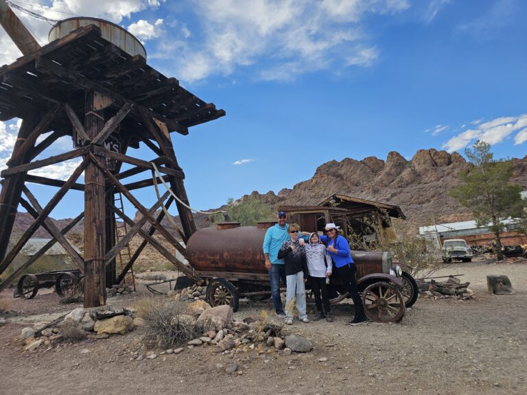 Smiling friends pose by a rusty truck and water tower in a Nevada ghost town near Las Vegas, perfect for ATV and RZR tours.