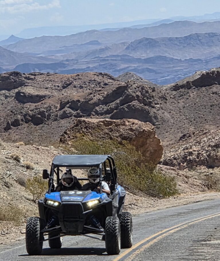 RZR off-road vehicle with two riders on a winding Nevada desert trail near Las Vegas and Colorado River under clear blue sky.