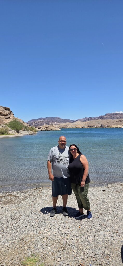 A smiling couple on a rocky Nevada shore by the Colorado River near Las Vegas, with blue skies and hills—great for ATV tours.