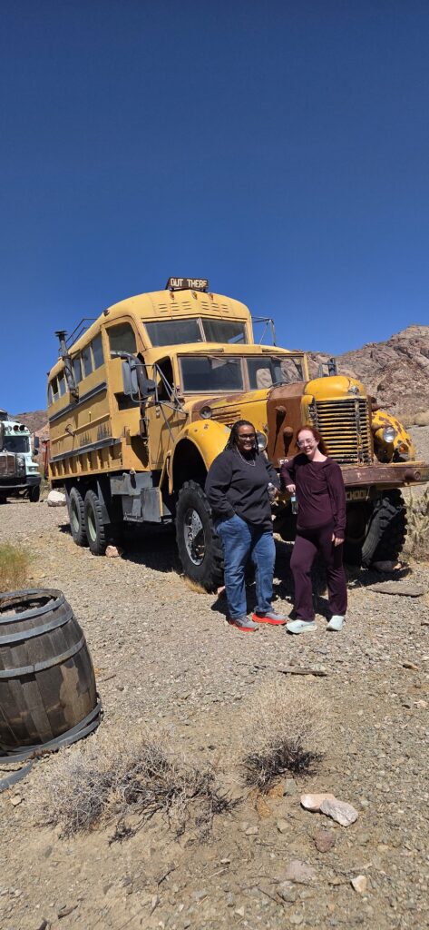 Smiling duo by a vintage yellow OUT THERE bus in Nevada, ready for Ghost Town sightseeing near the Colorado River and Las Vegas.