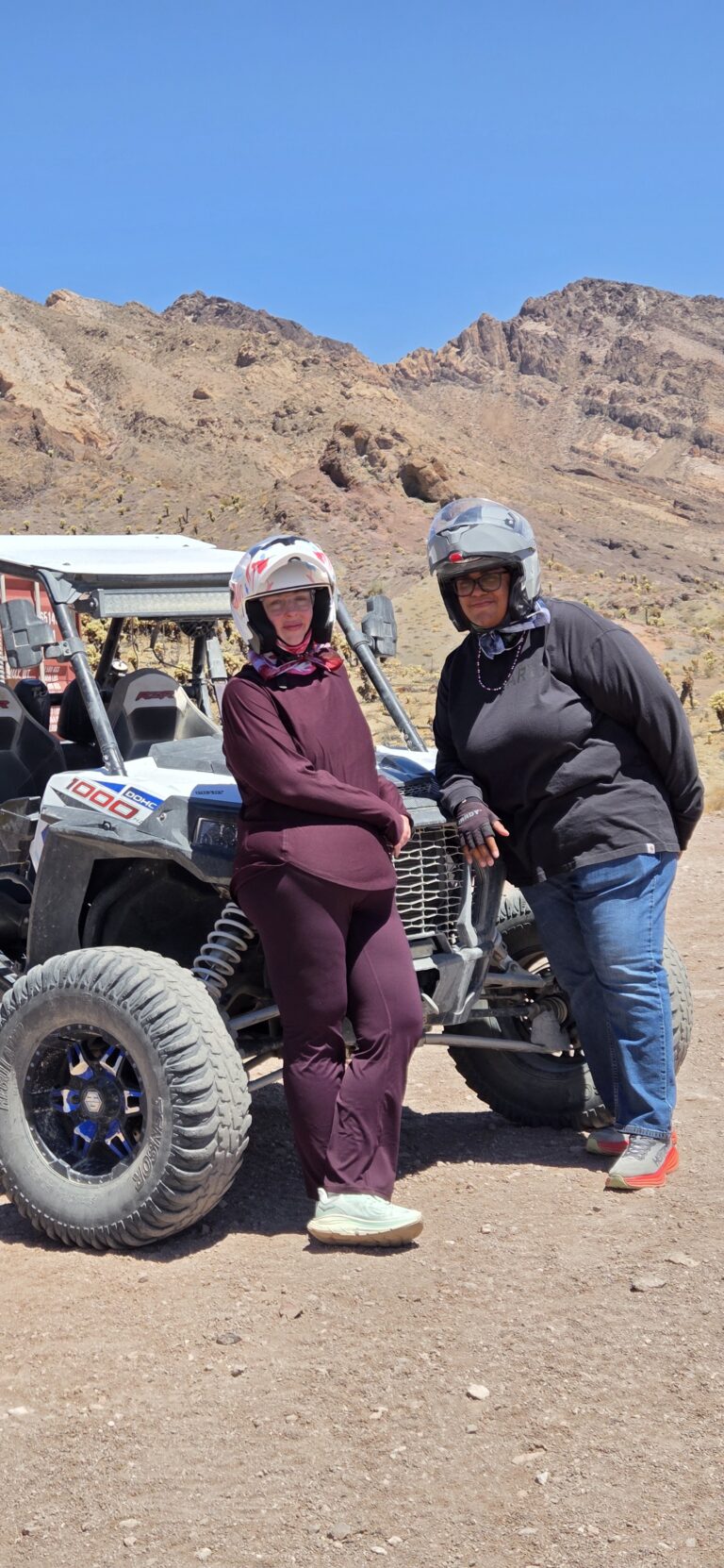 Two people in helmets pose by an off-road ATV in a rocky Nevada desert near Las Vegas with mountains and blue sky behind.