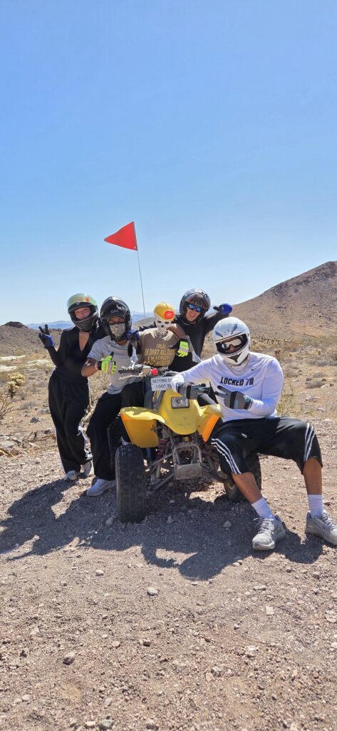Group in helmets poses with yellow ATV and red flag in Nevada desert near Las Vegas on a thrilling ATV tour under blue skies.
