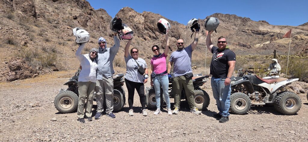 Group enjoying ATV tour in Nevada desert near Las Vegas, holding helmets with mountains and blue sky in the background.