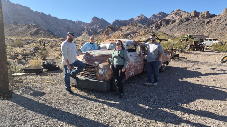 Four people by a rusty vintage truck in a Nevada desert ghost town near the Colorado River, with abandoned vehicles nearby.