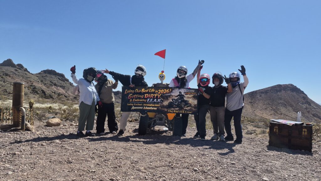 Seven adventurers in helmets pose on Nevada desert rocks near Las Vegas, waving a banner on an ATV tour under blue skies.