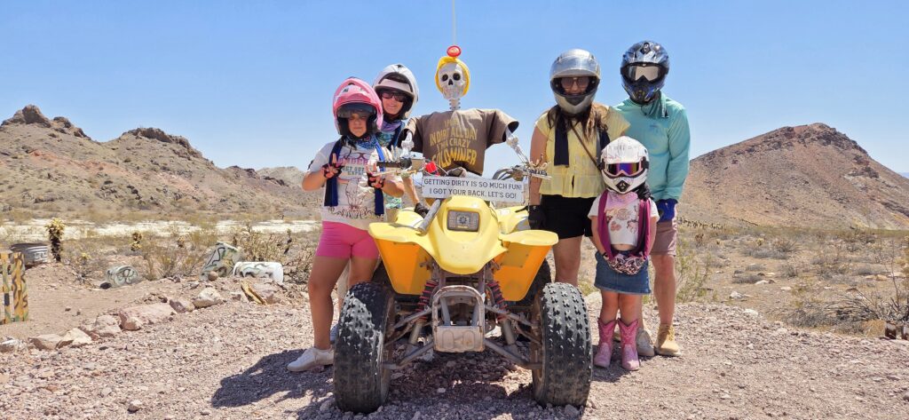 Group in helmets poses by yellow ATV with skeleton near Nevada desert, perfect for Las Vegas ghost town ATV tours and sightseeing.
