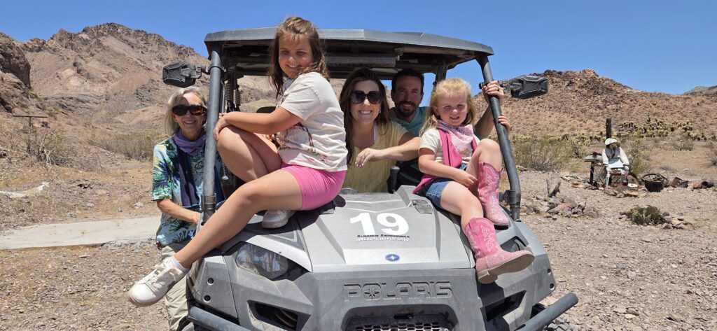 Family of five enjoys a Polaris RZR off-road ride near Las Vegas, Nevada desert, with rocky hills and blue skies in the background.