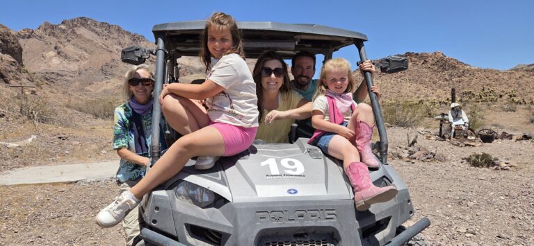 Family of five enjoys a Polaris RZR off-road ride near Las Vegas, Nevada desert, with rocky hills and blue skies in the background.