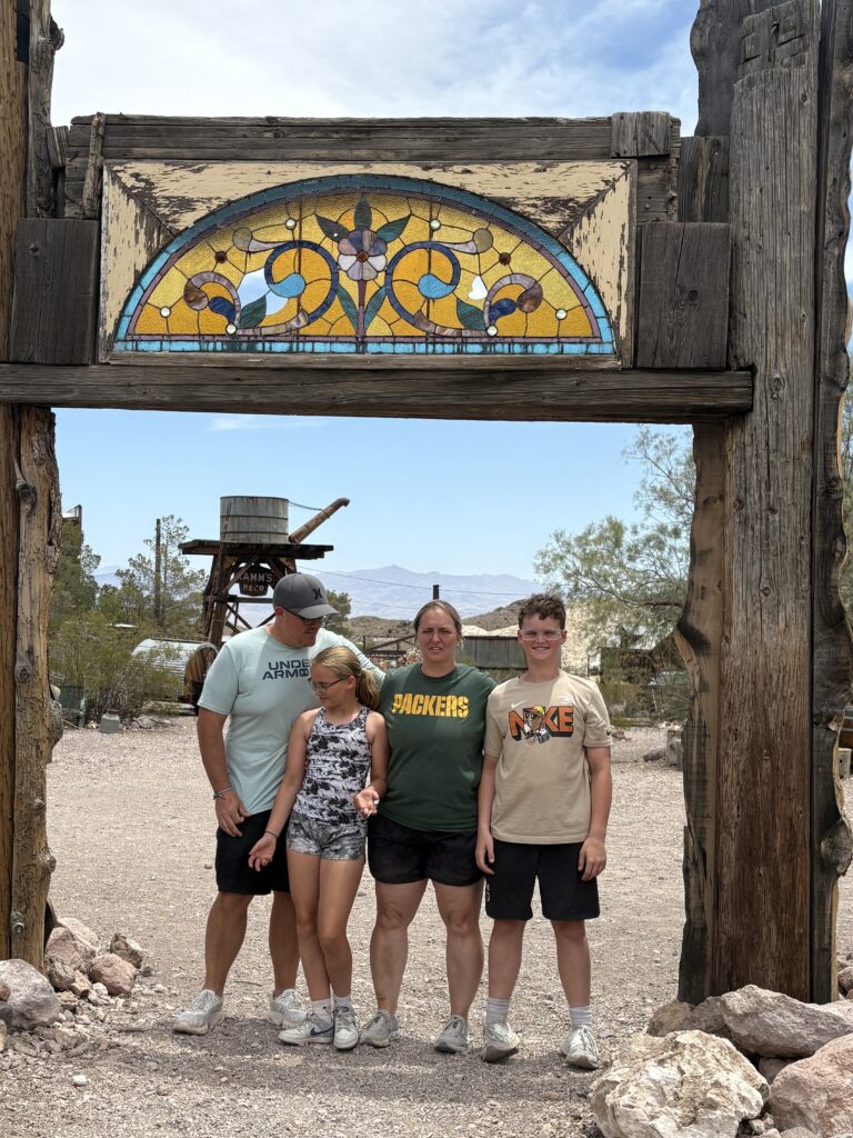Family of four beneath rustic wooden arch with stained glass near Nevada mountains, perfect for Las Vegas ghost town sightseeing.