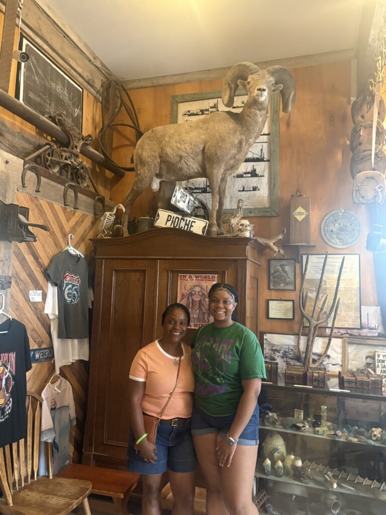 Two women pose in a rustic Nevada museum with taxidermy ram and antiques, perfect for Las Vegas Ghost Town sightseeing tours.