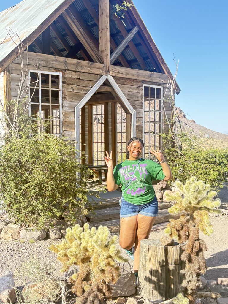 Smiling woman makes peace signs near rustic cabin with cacti in Nevada desert, perfect for Las Vegas ghost town sightseeing.