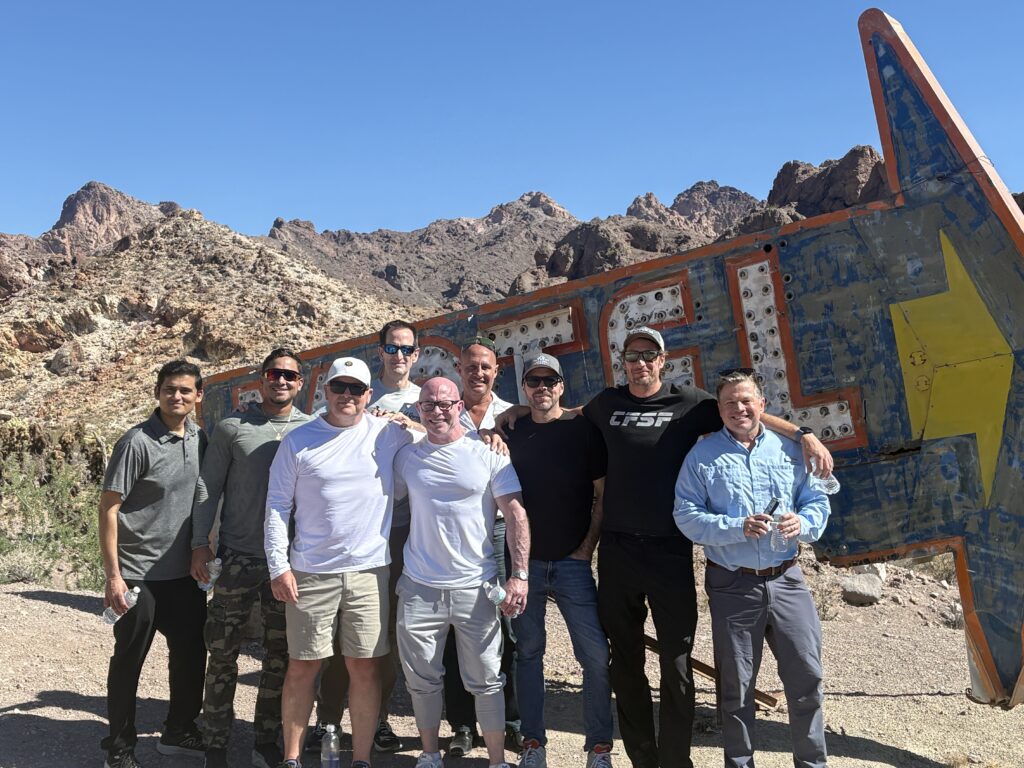 Nine men smile by a vintage sign with Nevada mountains, clear skies—perfect for Las Vegas ATV tours or Ghost Town sightseeing.