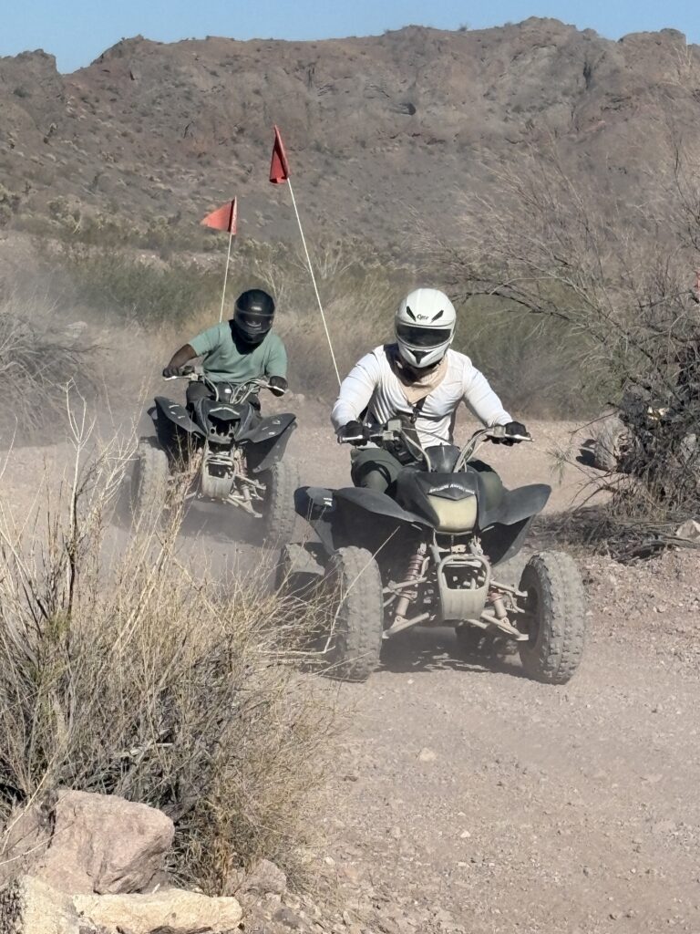 ATV riders with helmets blaze a dusty Nevada desert trail near Las Vegas, red flags flying by rocky hills—perfect for ATV tours.