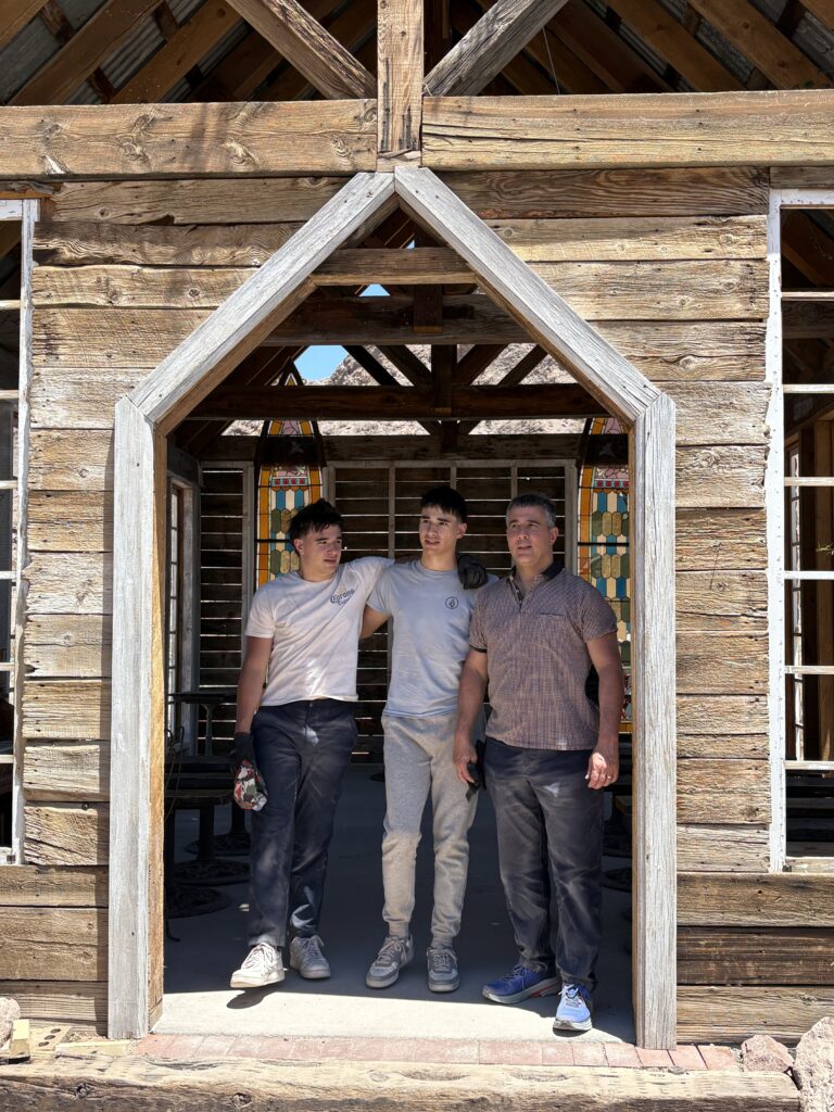 Three men stand in a rustic Nevada ghost town doorway near the Colorado River, sunlight streaming through exposed wooden beams.