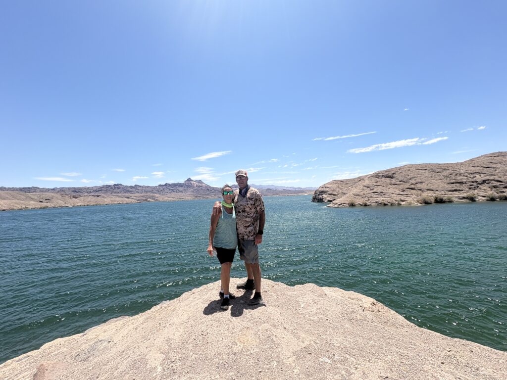 Two people on a rocky outcrop near the Colorado River, overlooking a blue lake in Nevada’s arid mountains under a sunny sky.