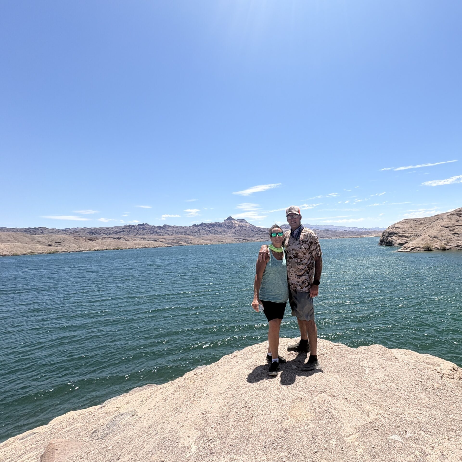 Two people on a rocky outcrop near the Colorado River, overlooking a blue lake in Nevada’s arid mountains under a sunny sky.