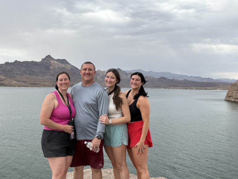 Four friends in athletic wear smile by a Nevada lake, mountains behind them, after an ATV tour near the Colorado River, Las Vegas.