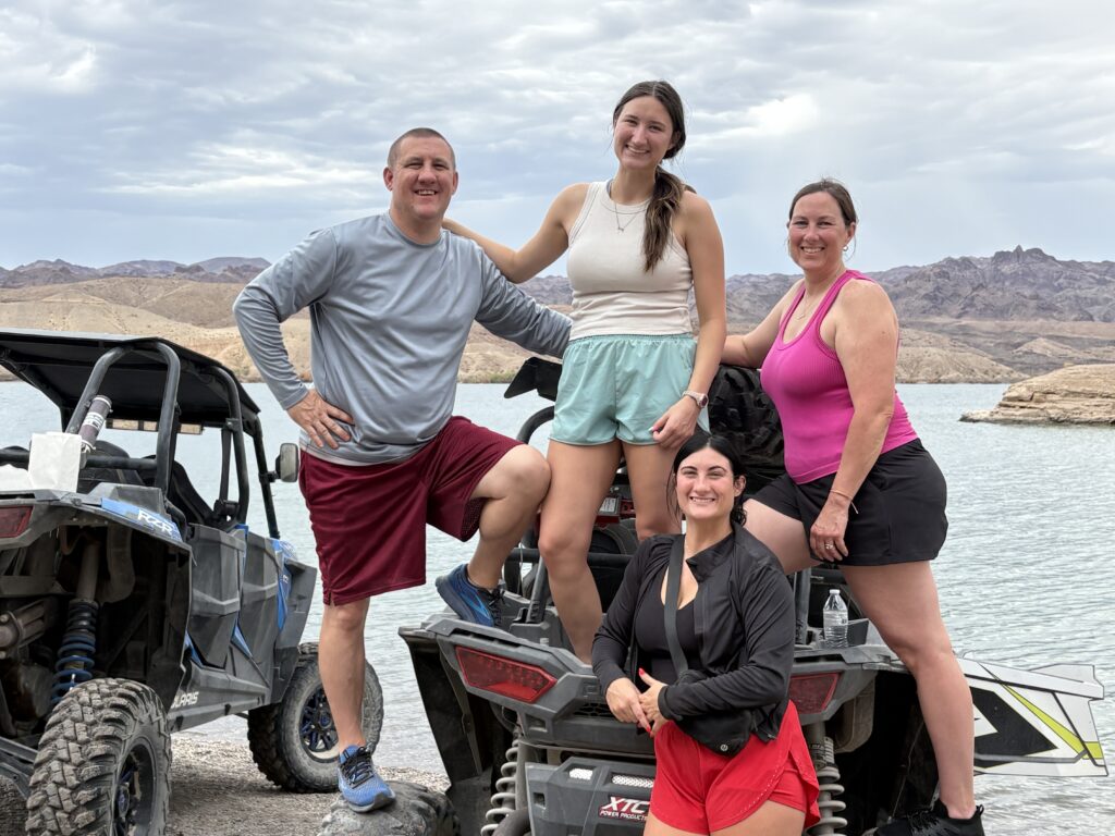 Four friends in athletic wear smile on ATVs by a lake near the Colorado River, Nevada, enjoying a scenic RZR off-road tour.