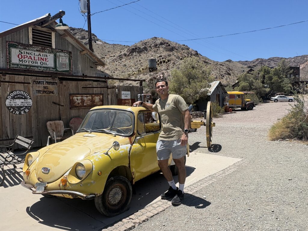 Smiling man by yellow vintage car at Nevada ghost town near Las Vegas, rustic signs, perfect for sightseeing or ATV tours.