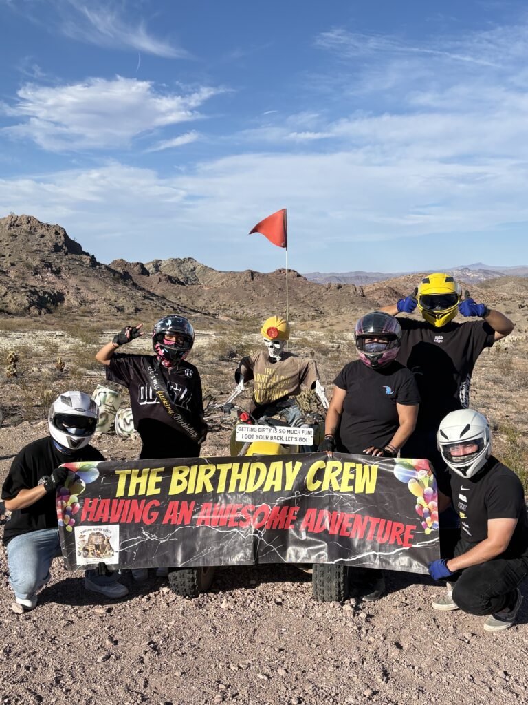 Five helmeted friends in Nevada desert with birthday banner, ready for an ATV tour near Las Vegas and Colorado River.