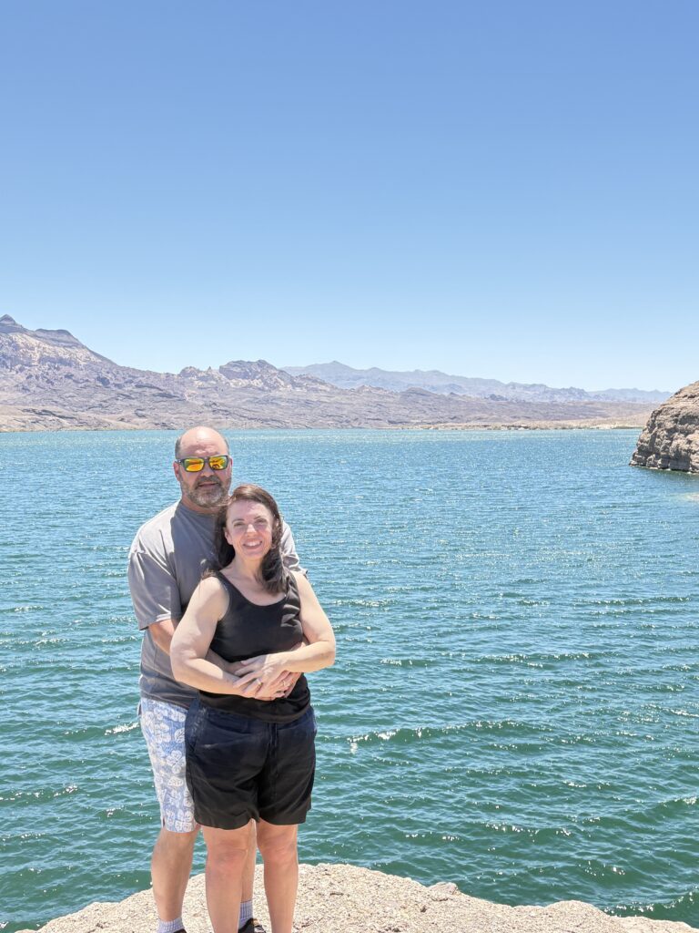 Smiling couple on rocky Nevada shore by the bright blue Colorado River, mountains behind, perfect for Las Vegas ATV tours.