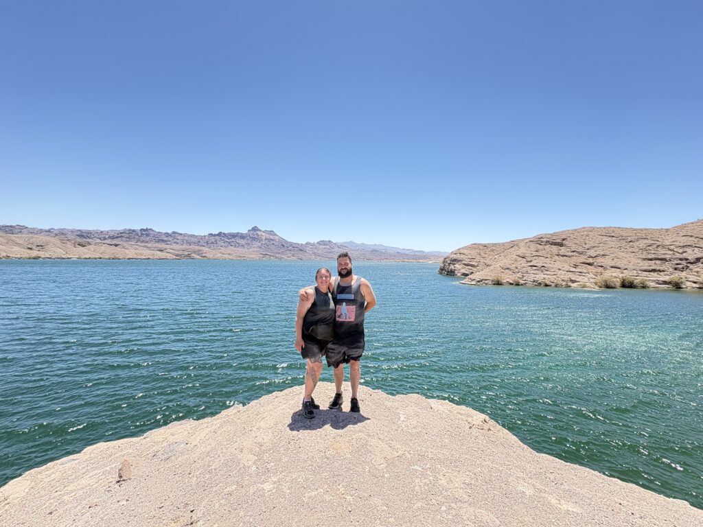 Two people on a rocky outcrop overlook the blue Colorado River near Las Vegas, Nevada, with arid hills and clear skies behind.