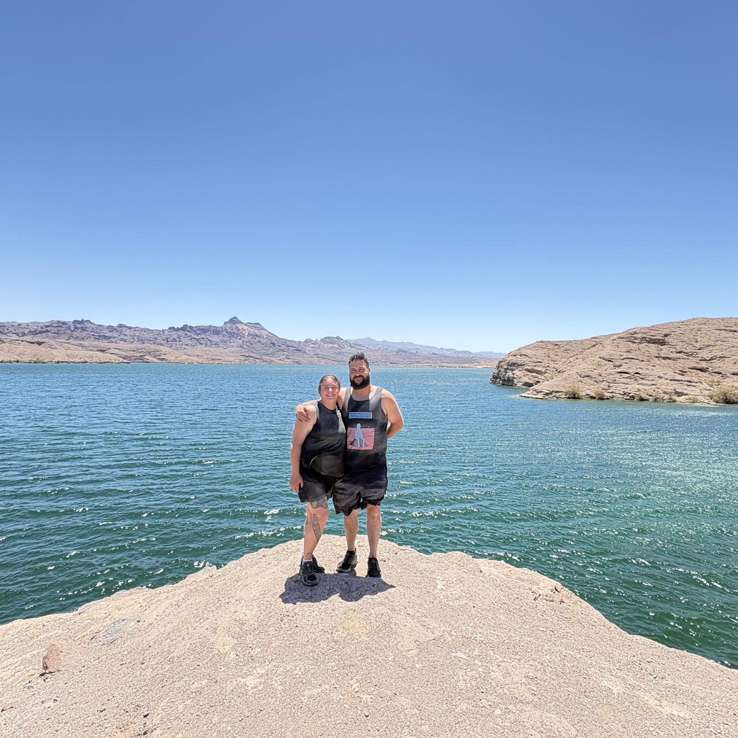 Two people on a rocky outcrop overlook the blue Colorado River near Las Vegas, Nevada, with arid hills and clear skies behind.