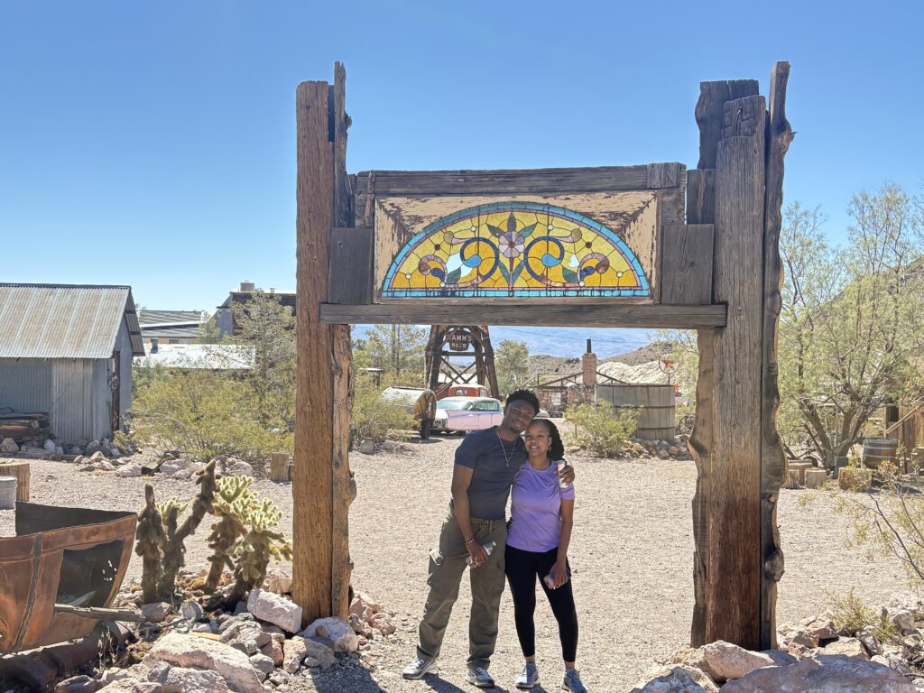 Smiling duo under a stained glass arch in a Nevada ghost town near Las Vegas, with cacti, vintage car & mining gear—perfect for sightseeing.