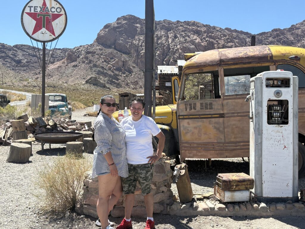 Smiling friends at a vintage gas pump and yellow bus in a Nevada ghost town near Las Vegas, perfect for sightseeing tours.