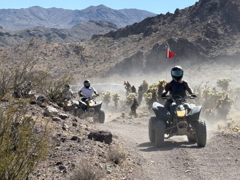 Adventurers on ATVs tour a dusty Nevada desert trail near Las Vegas, surrounded by rocky hills, cacti, and distant mountains.
