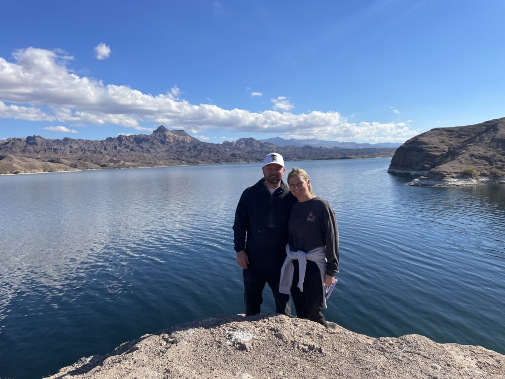 Smiling couple on a rocky ledge by Nevada’s Colorado River near Las Vegas, scenic mountains, and blue sky in the background.