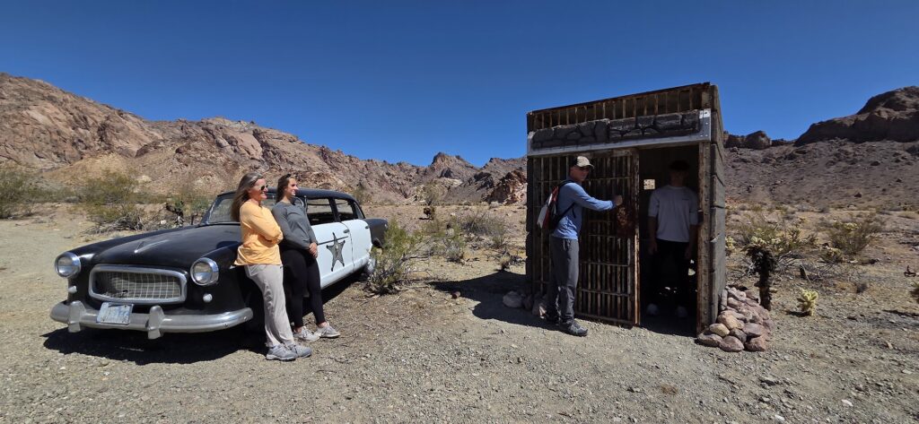 Tourists near a vintage car in a Nevada desert ghost town, sightseeing by a rustic jail with mountains near Las Vegas.