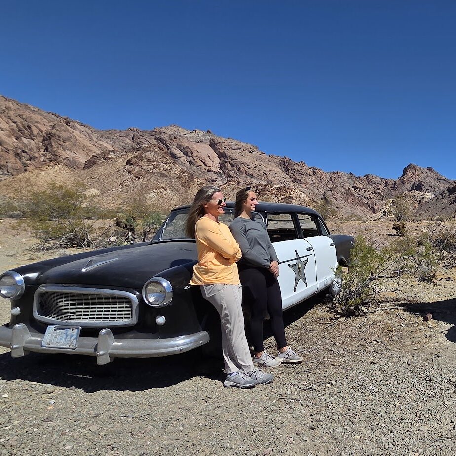 Tourists near a vintage car in a Nevada desert ghost town, sightseeing by a rustic jail with mountains near Las Vegas.