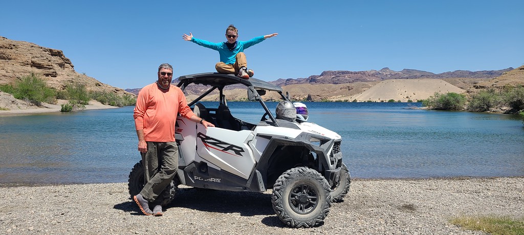 Family enjoys off-road adventure by Colorado River near Las Vegas, Nevada—child on vehicle roof, scenic rocky hills in background.
