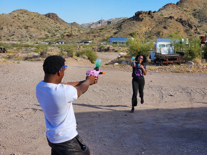 Smiling duo with colorful water guns in a Nevada desert near Las Vegas, old vehicles and hills, enjoying off-road adventure fun.
