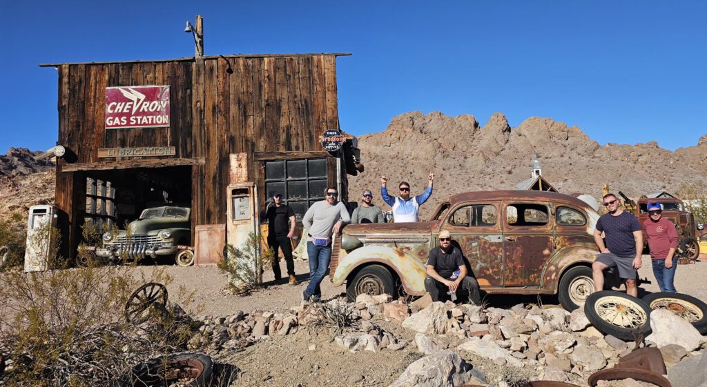 Seven friends smile by a rusty vintage car at Nelson Ghost Town near Las Vegas, Nevada—perfect for Ghost Town sightseeing tours.
