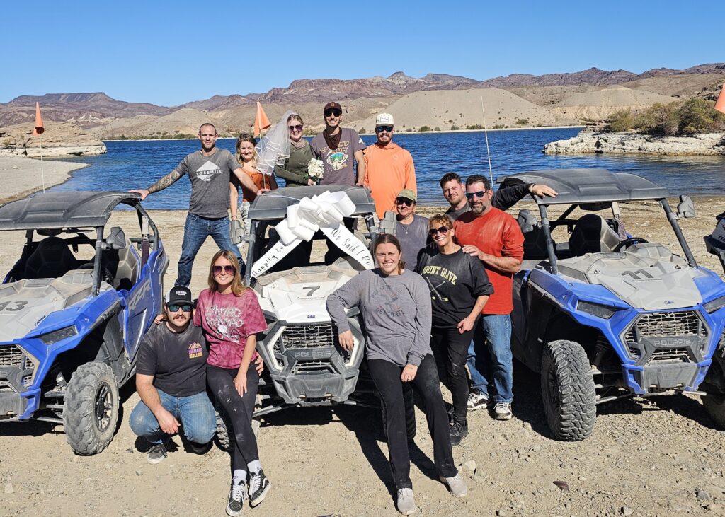 Group smiles by ATVs with a white bow on sandy Nevada lakeshore near Colorado River, Las Vegas. RZR off-road adventure.