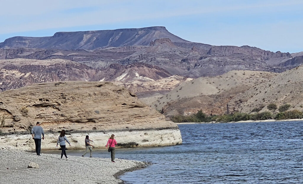 Four people explore the rocky Colorado River shoreline near Las Vegas, Nevada, with scenic mountains and sandstone formations.